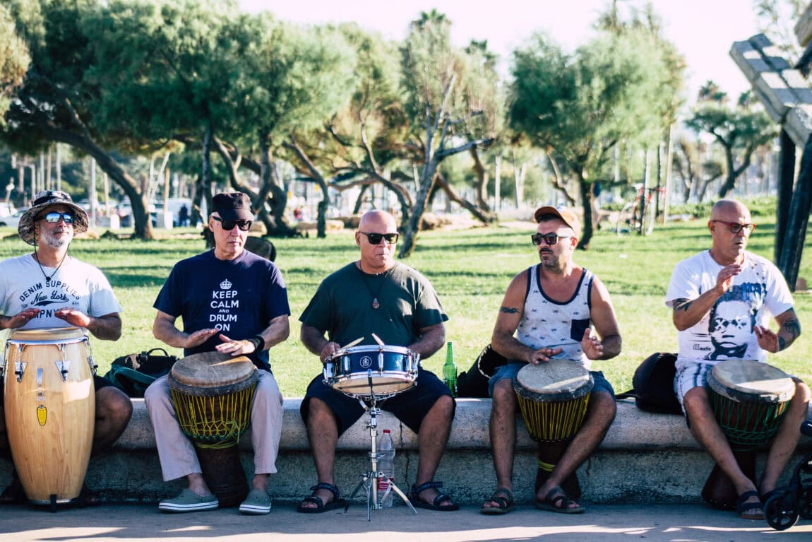 People playing drums outdoors in a park, enjoying a sunny day, with trees and green space in the background. A group of five men seated on a bench, each with different types of drums, engaged in music and rhythm.