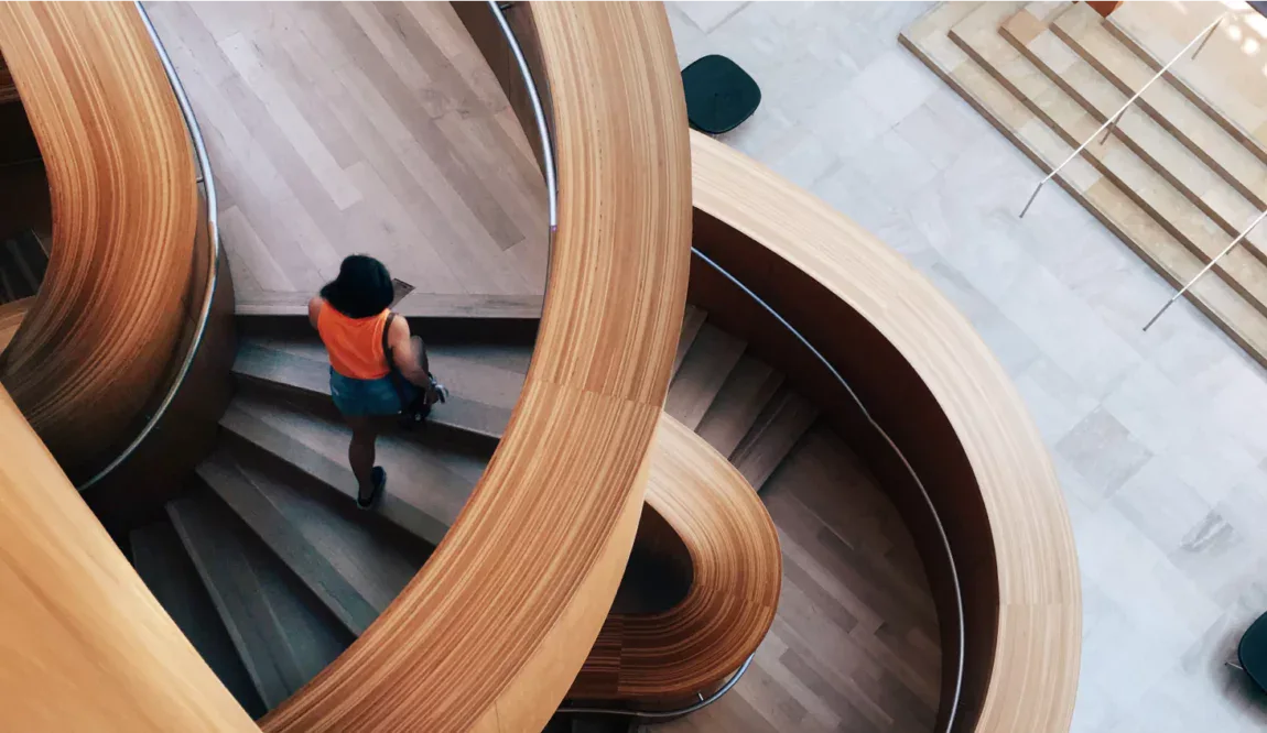Modern wooden spiral staircase with a person walking up, featuring smooth curves and sleek design, in a contemporary interior space.